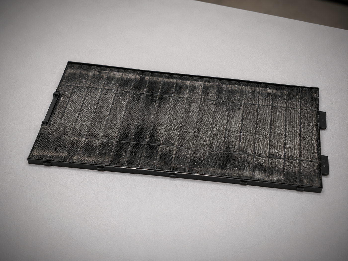 Dirty PC dust filter resting on a light gray table, showing heavy gray dust buildup across the mesh surface before cleaning.